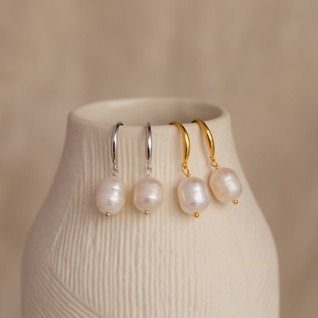 Two pairs of Dangling Pearl Drop Earrings—one in silver and one in gold—displayed hanging over the edge of a textured cream-colored ceramic vase.