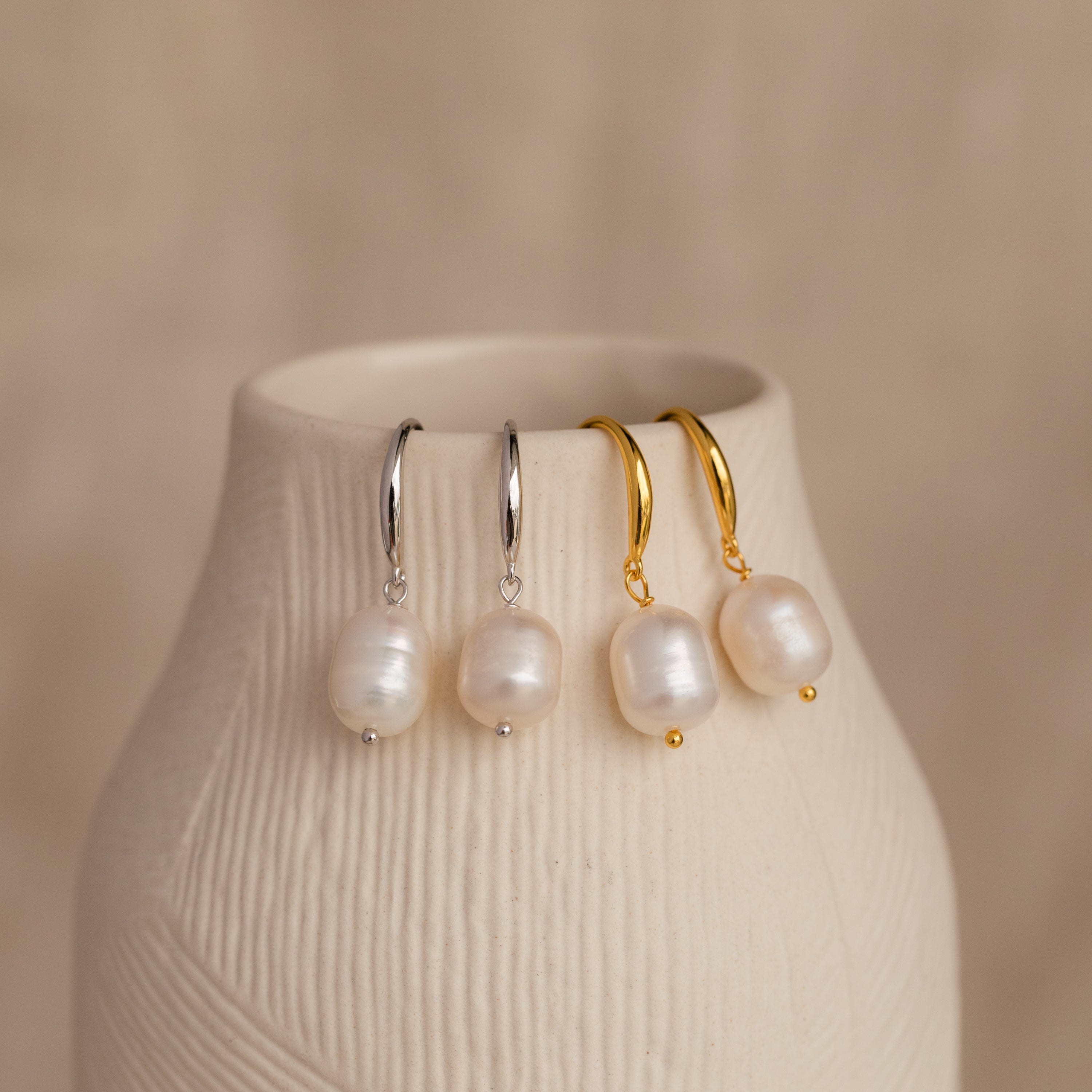 Two pairs of Dangling Pearl Drop Earrings—one in silver and one in gold—displayed hanging over the edge of a textured cream-colored ceramic vase.