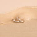 A close-up image of a pair of delicate silver huggie earrings, resting on a soft beige fabric. Each earring features a round white pearl set between two sparkling white gemstones.