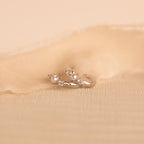 A close-up image of a pair of delicate silver huggie earrings, resting on a soft beige fabric. Each earring features a round white pearl set between two sparkling white gemstones.