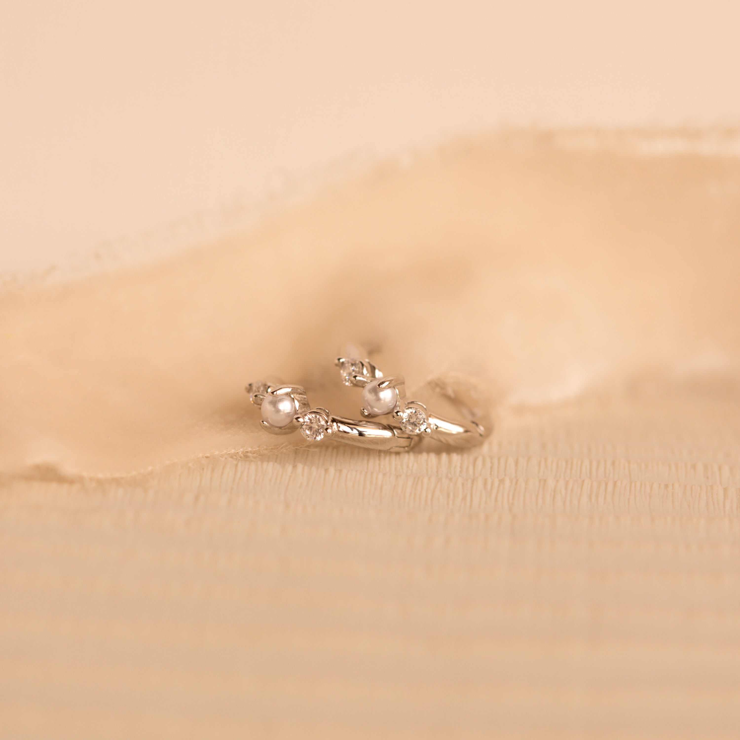 A close-up image of a pair of delicate silver huggie earrings, resting on a soft beige fabric. Each earring features a round white pearl set between two sparkling white gemstones.