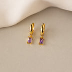 Pair of gold huggie hoop earrings with rectangular baguette-cut amethyst charms, displayed on a neutral surface with a soft cream fabric backdrop.