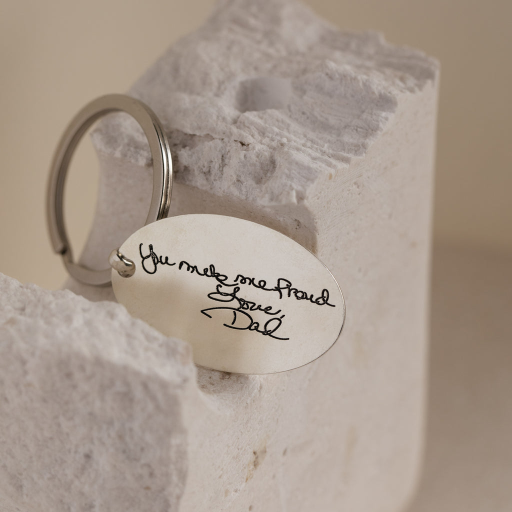 The Handwriting Oval Keychain features a custom engraving of a handwritten note—"You make me proud. Love, Dad"—and is beautifully displayed on a stone block.