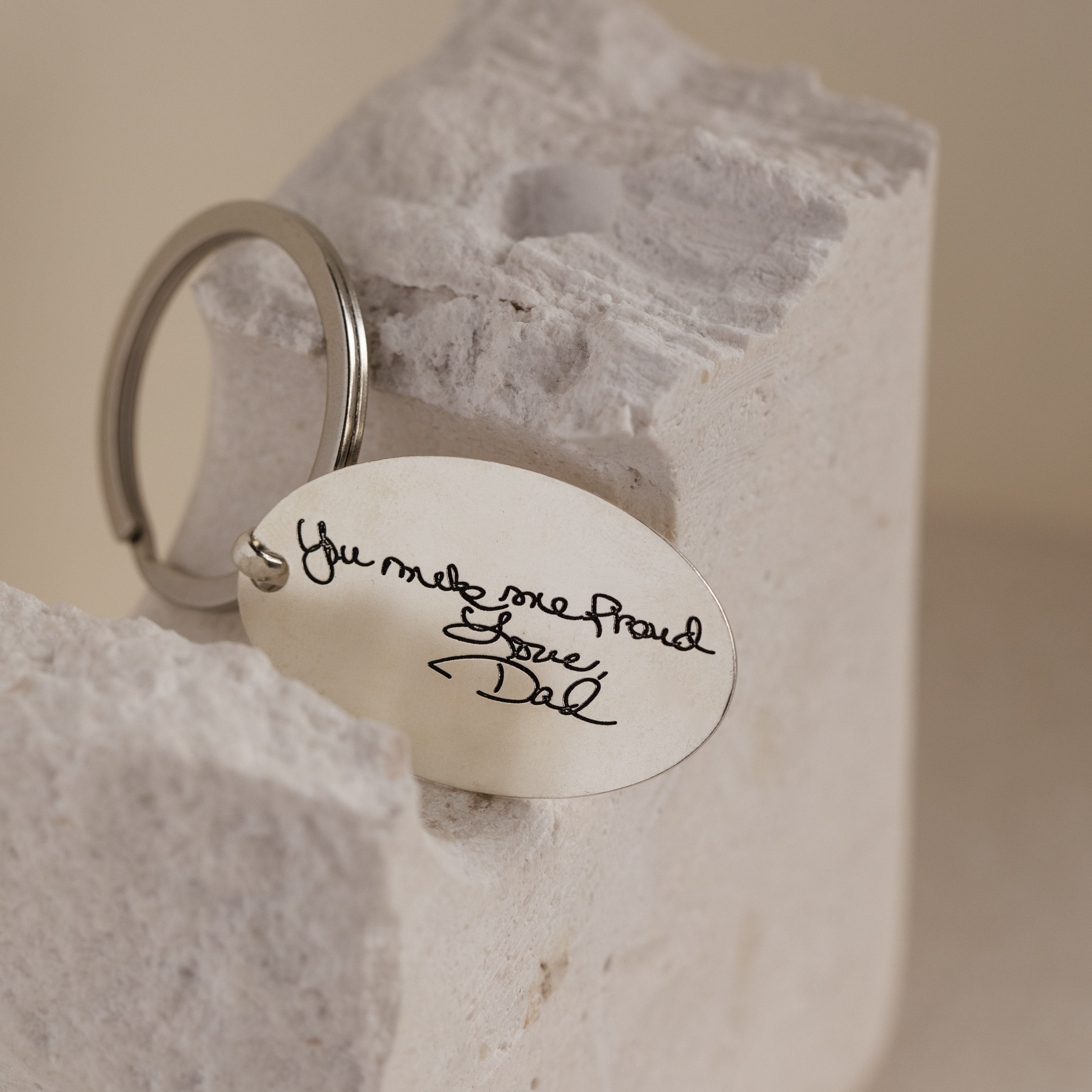 The Handwriting Oval Keychain features a custom engraving of a handwritten note—"You make me proud. Love, Dad"—and is beautifully displayed on a stone block.