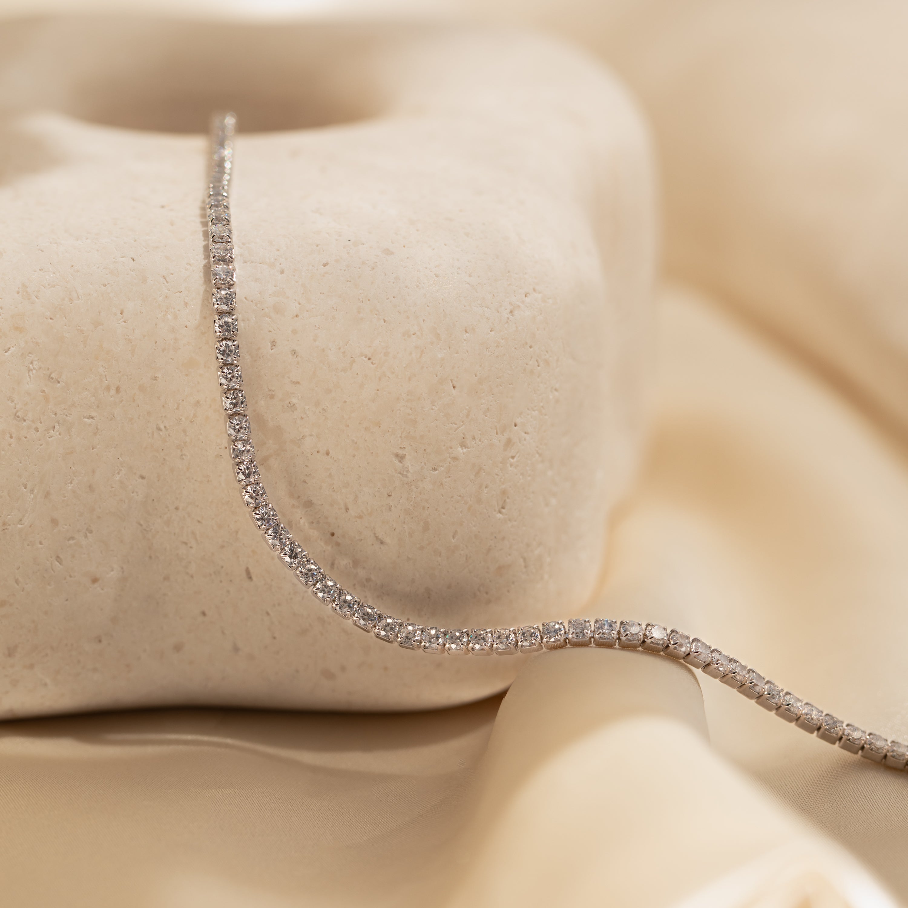 The Classic Diamond Tennis Bracelet in White Gold is elegantly displayed on textured stone with a soft beige satin background.