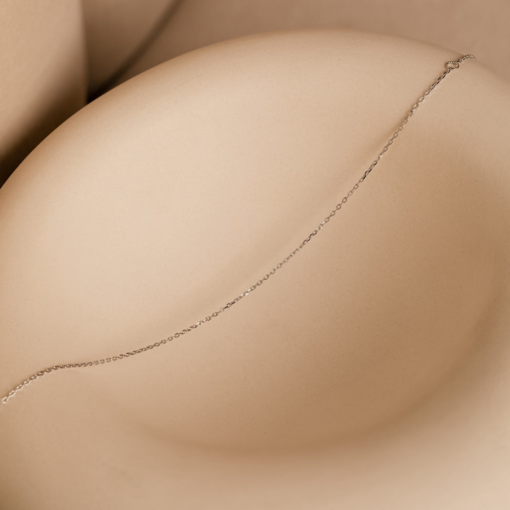 The Micro Rolo Chain Anklet in Sterling Silver is displayed on a smooth beige curved surface, its delicate silver chain highlighted by soft shadows.