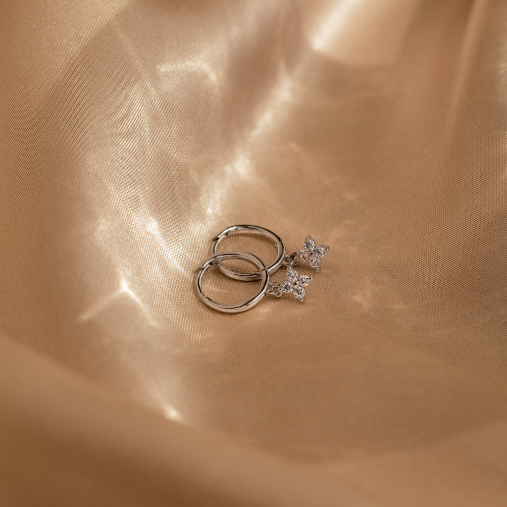 Silver Diamond Flower Huggies displayed on shimmery beige satin fabric. The hoops have a smooth, polished finish, while the flower charms are encrusted with sparkling clear stones. 
