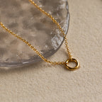 The Rolo Circle Charm Necklace features a simple circular pendant resting on a textured, transparent surface—an elegant and minimalist accessory perfect for everyday wear.