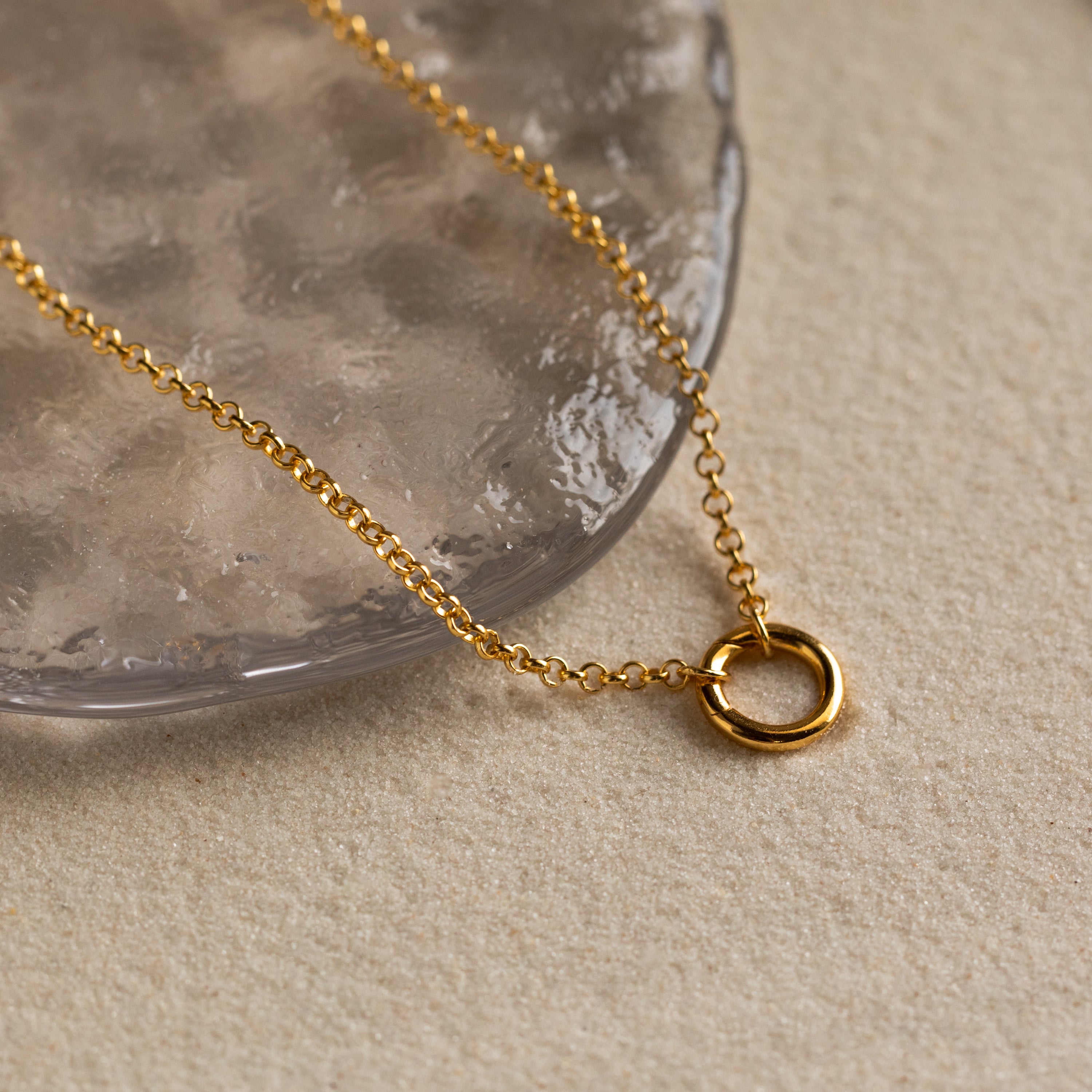 The Rolo Circle Charm Necklace features a simple circular pendant resting on a textured, transparent surface—an elegant and minimalist accessory perfect for everyday wear.