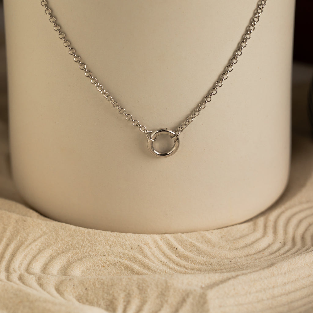 The Rolo Circle Charm Necklace in Sterling Silver features a small circular pendant on a sleek chain, displayed on a sand-patterned neutral background. This customizable piece brings subtle elegance to any outfit.