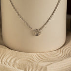 The Rolo Circle Charm Necklace in Sterling Silver features a small circular pendant on a sleek chain, displayed on a sand-patterned neutral background. This customizable piece brings subtle elegance to any outfit.