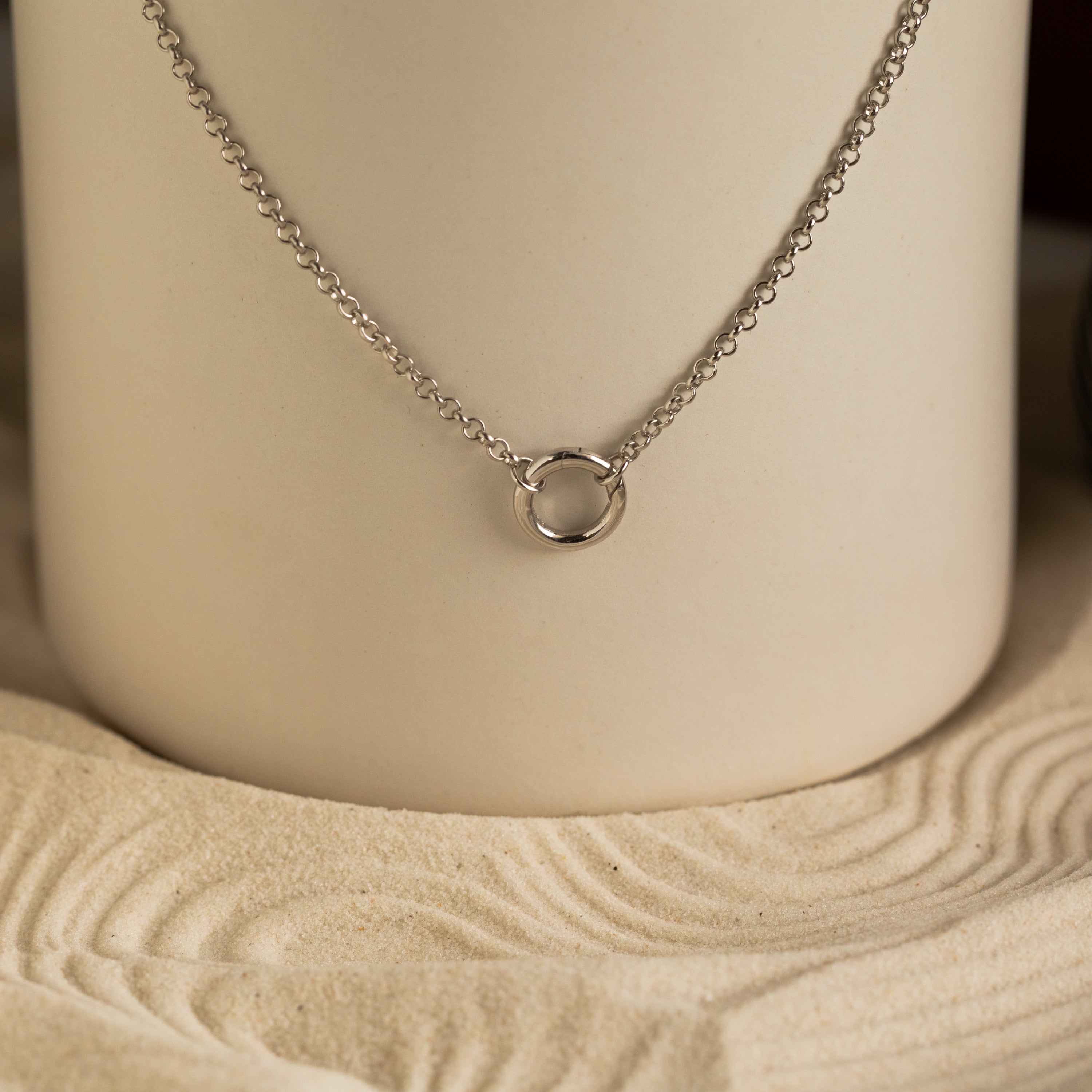 The Rolo Circle Charm Necklace in Sterling Silver features a small circular pendant on a sleek chain, displayed on a sand-patterned neutral background. This customizable piece brings subtle elegance to any outfit.