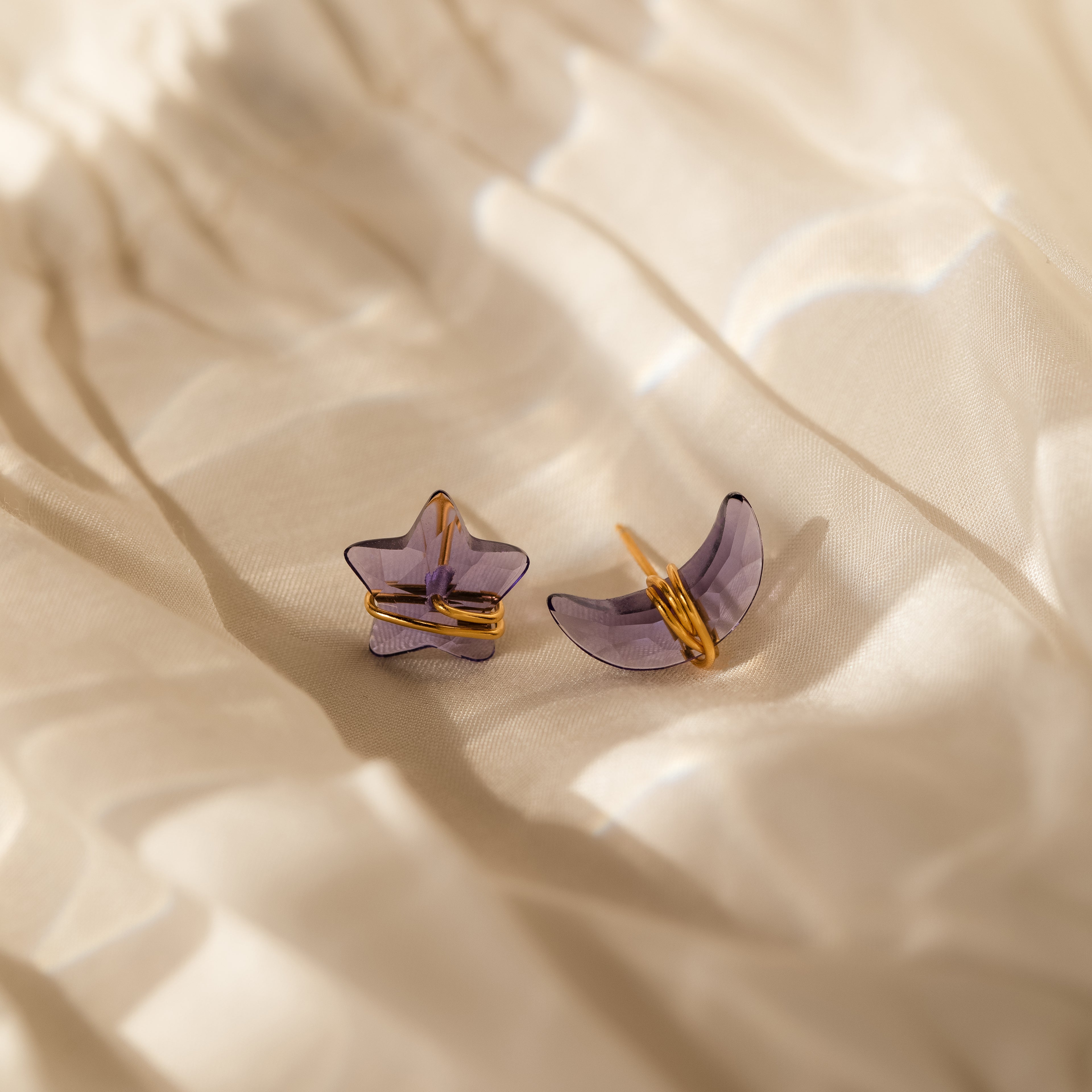 Close up of a pair of purple amethyst moon and star-shaped earrings in gold resting on a beige fabric.