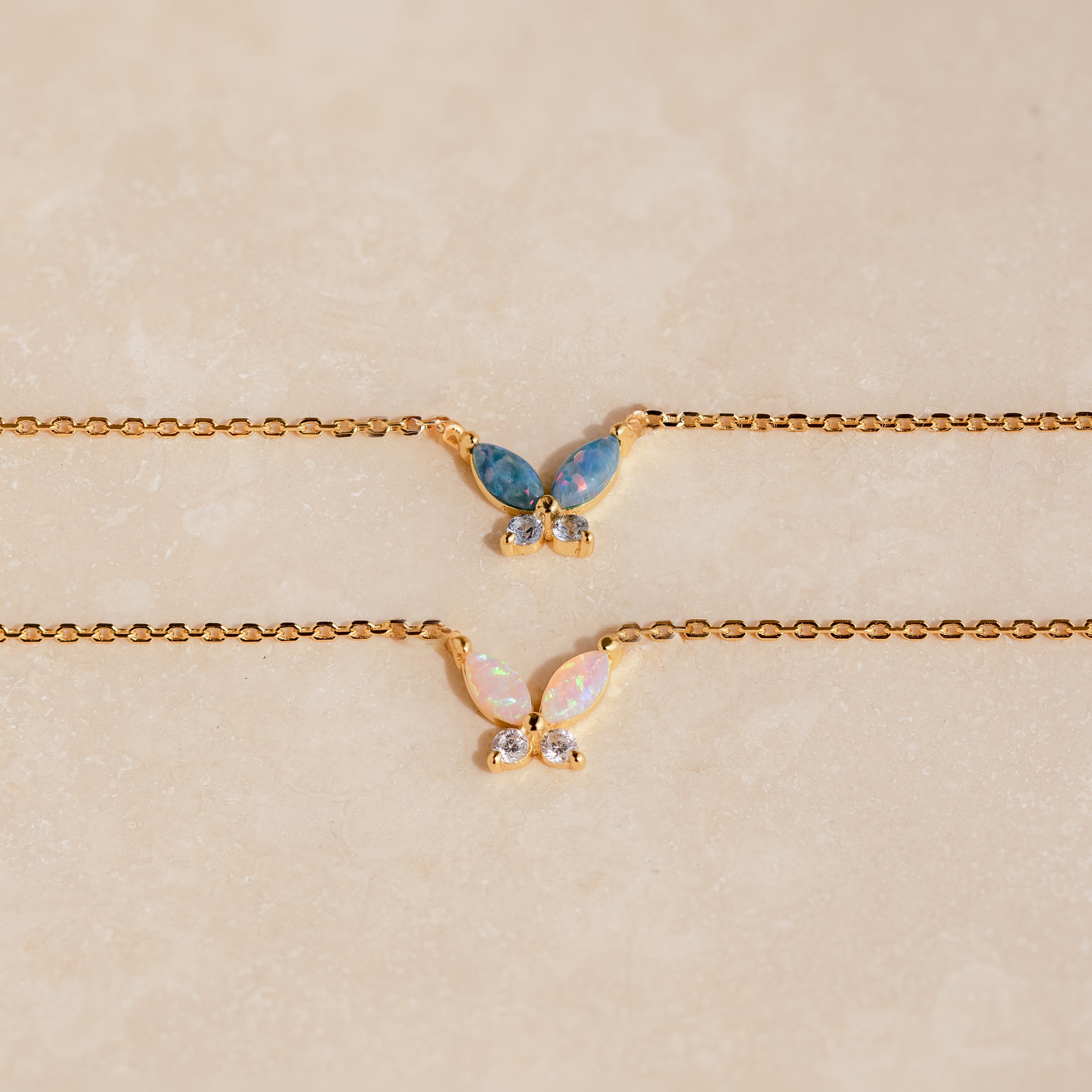 Two gold butterfly necklaces with opal wings, one in blue opal and the other in white opal, accented with small clear stones.
