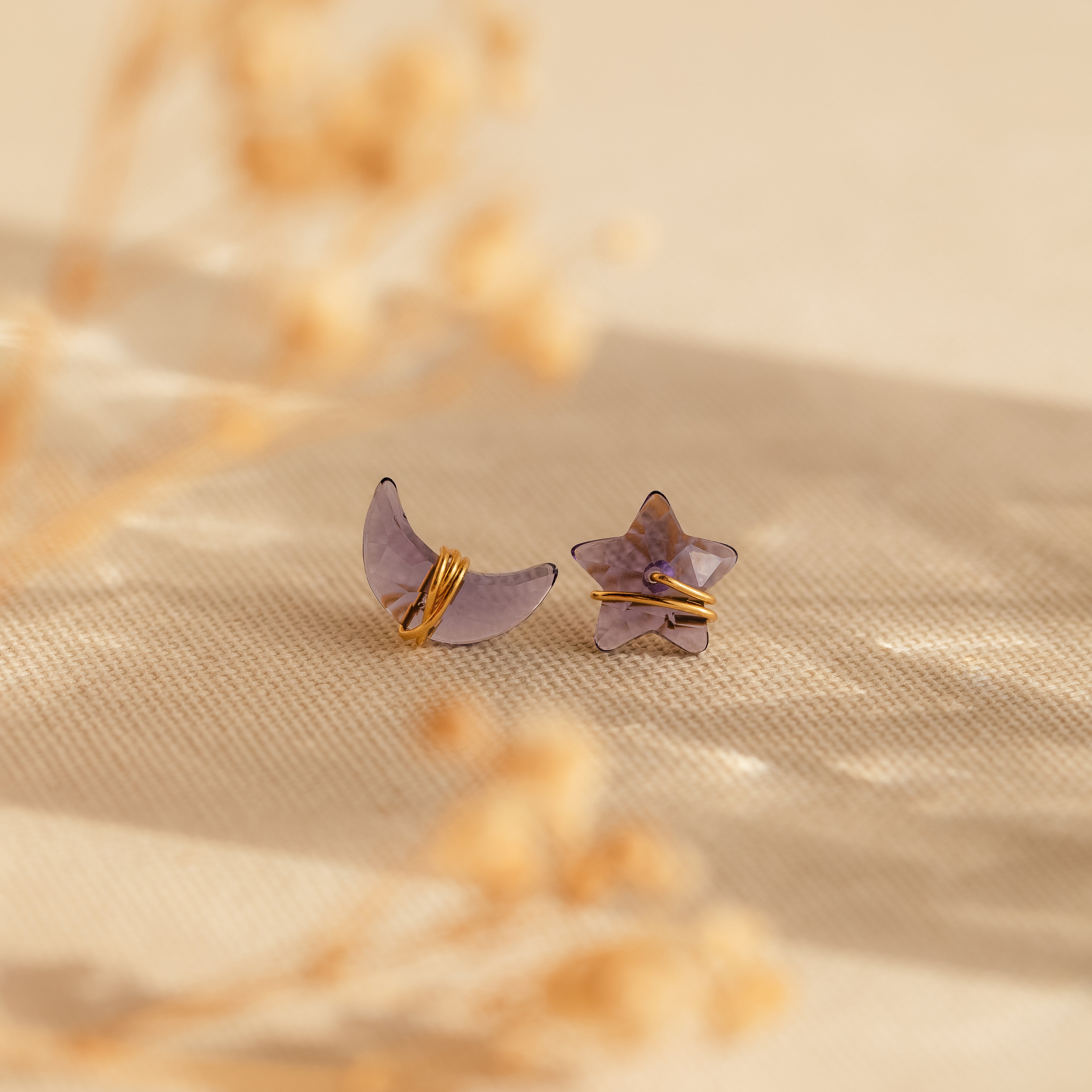 Purple moon and star earrings in gold sit on a beige fabric with dried flowers in the background.