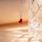 The Little Red Heart Necklace, a silver minimalist piece with a red heart pendant, is displayed beside a clear faceted glass object.