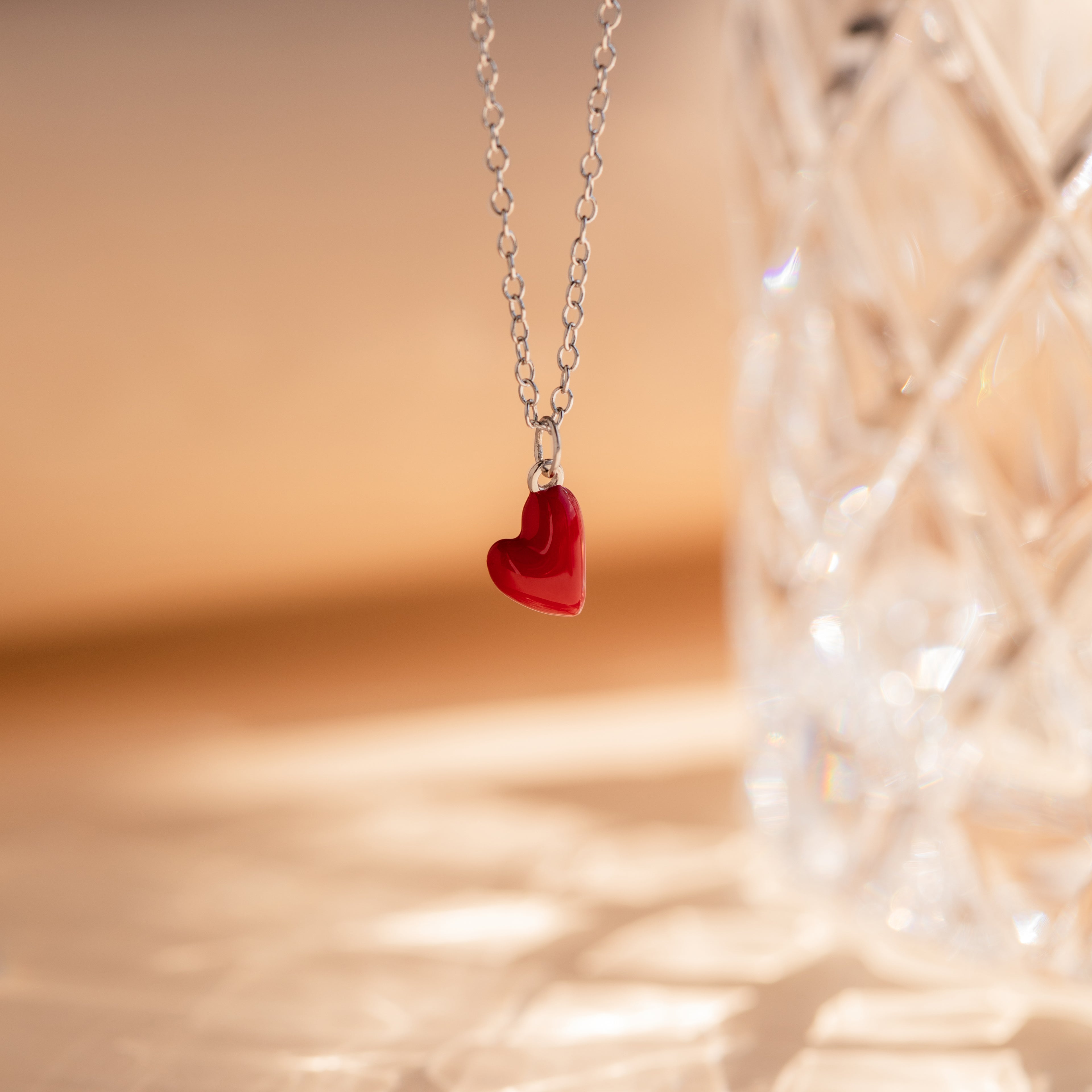 The Little Red Heart Necklace, a silver minimalist piece with a red heart pendant, is displayed beside a clear faceted glass object.