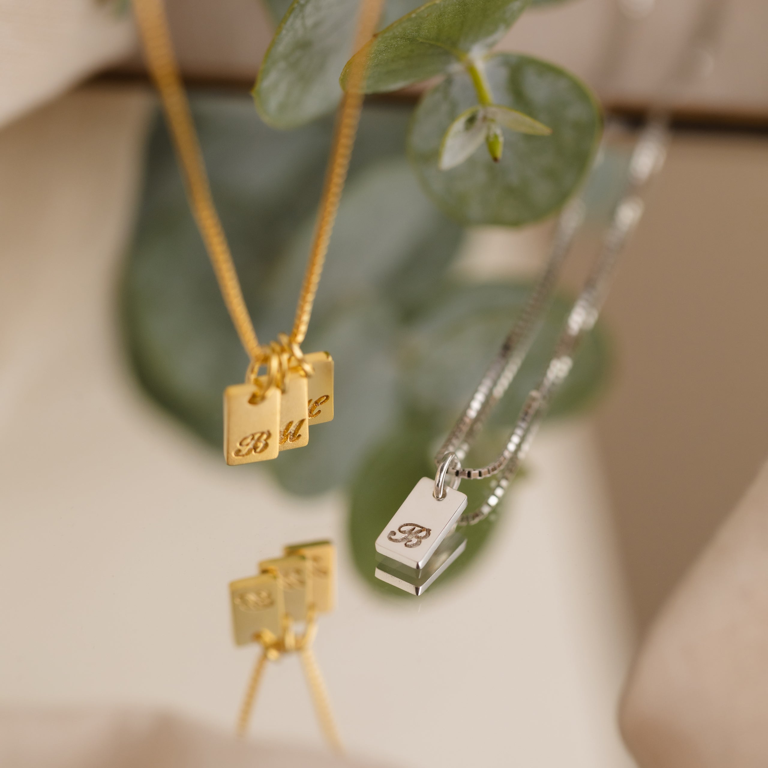 Gold and silver minimalist necklaces with engraved rectangular pendants reflect on a surface, green leaves in the background. Discover the Carrie Mini Tag Necklace for a subtle, personalized touch.
