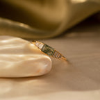 Close-up of a gold ring featuring a rectangular green moss agate gemstone at the center, flanked by three baguette-cut white stones on each side, resting elegantly on a smooth, light-toned stone surface with a golden satin backdrop.