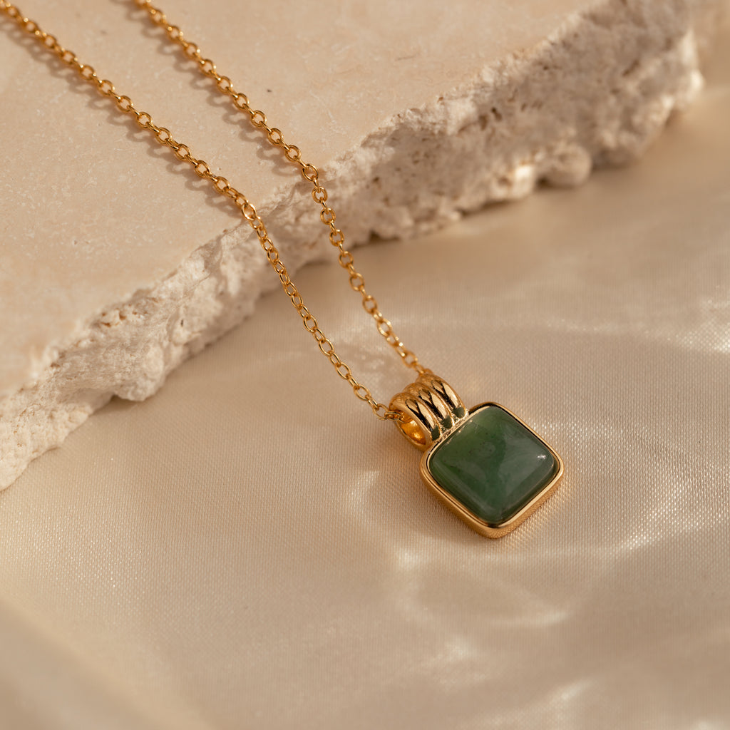 The Jade Pendant Necklace in 18K Gold features a square green jade stone, symbolizing luck, and is showcased on a textured beige surface.