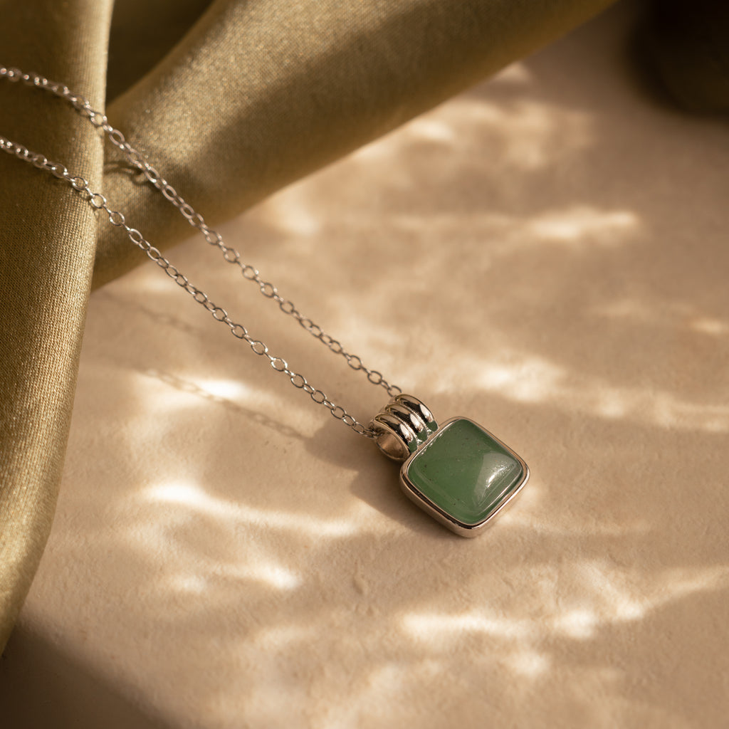 The Jade Pendant Necklace in Silver features a square green jade stone resting on a beige surface under soft, dappled light—symbolizing luck and elegance.