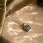 The Jade Pendant Necklace in Silver features a square green jade stone resting on a beige surface under soft, dappled light—symbolizing luck and elegance.