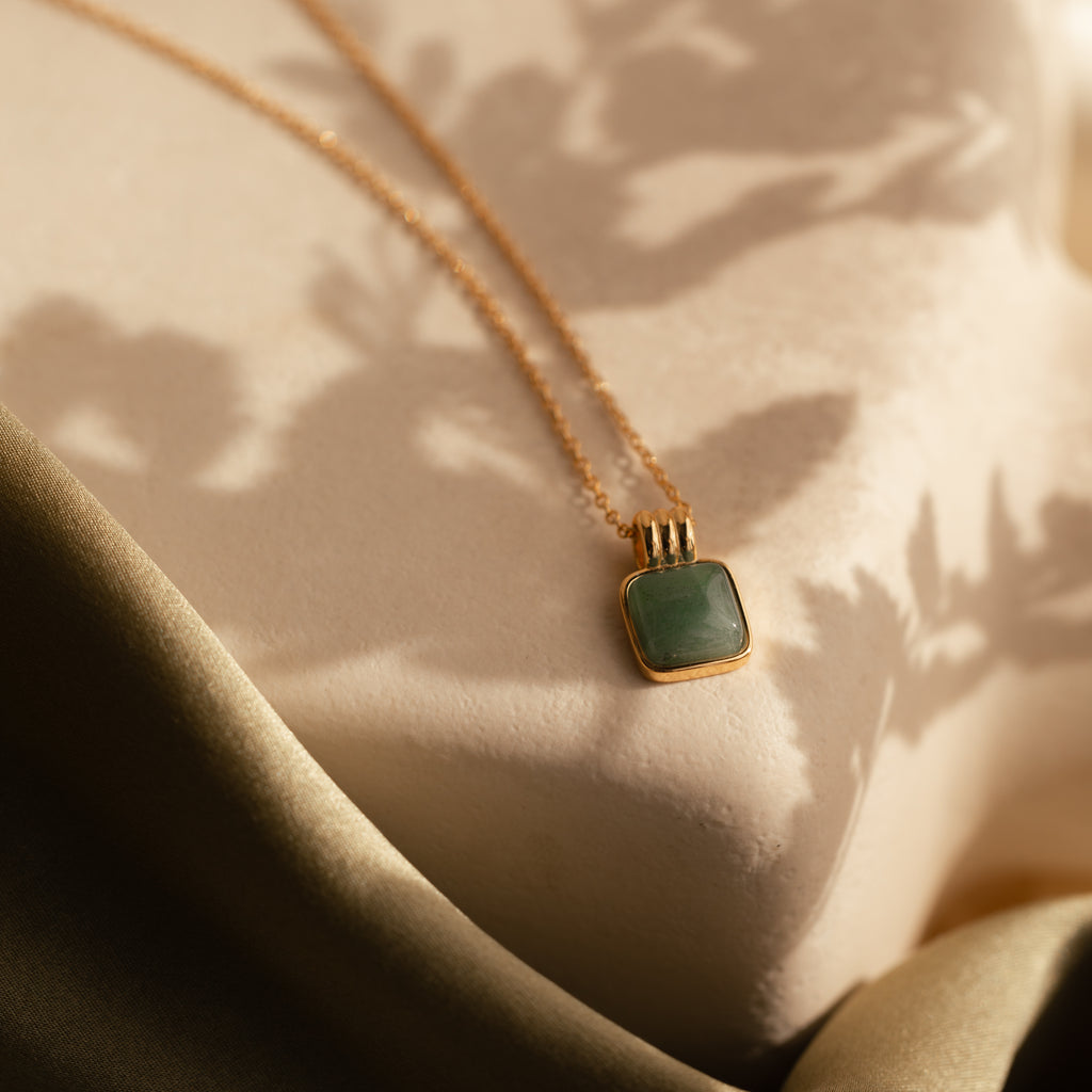 The Jade Pendant Necklace with a green jade stone rests on a soft beige surface with leafy shadows and fabric—perfect for symbolizing new beginnings.