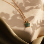 The Jade Pendant Necklace with a green jade stone rests on a soft beige surface with leafy shadows and fabric—perfect for symbolizing new beginnings.