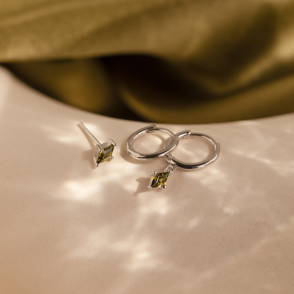 The Peridot Earrings Set in Sterling Silver features silver hoops and green gemstone studs for a chic stacking look, displayed on a light textured surface.