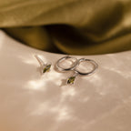The Peridot Earrings Set in Sterling Silver features silver hoops and green gemstone studs for a chic stacking look, displayed on a light textured surface.