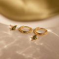The Peridot Earrings Set in 18K Gold features gold hoop stacking earrings with green gemstone charms, beautifully displayed on a creamy, softly lit surface.