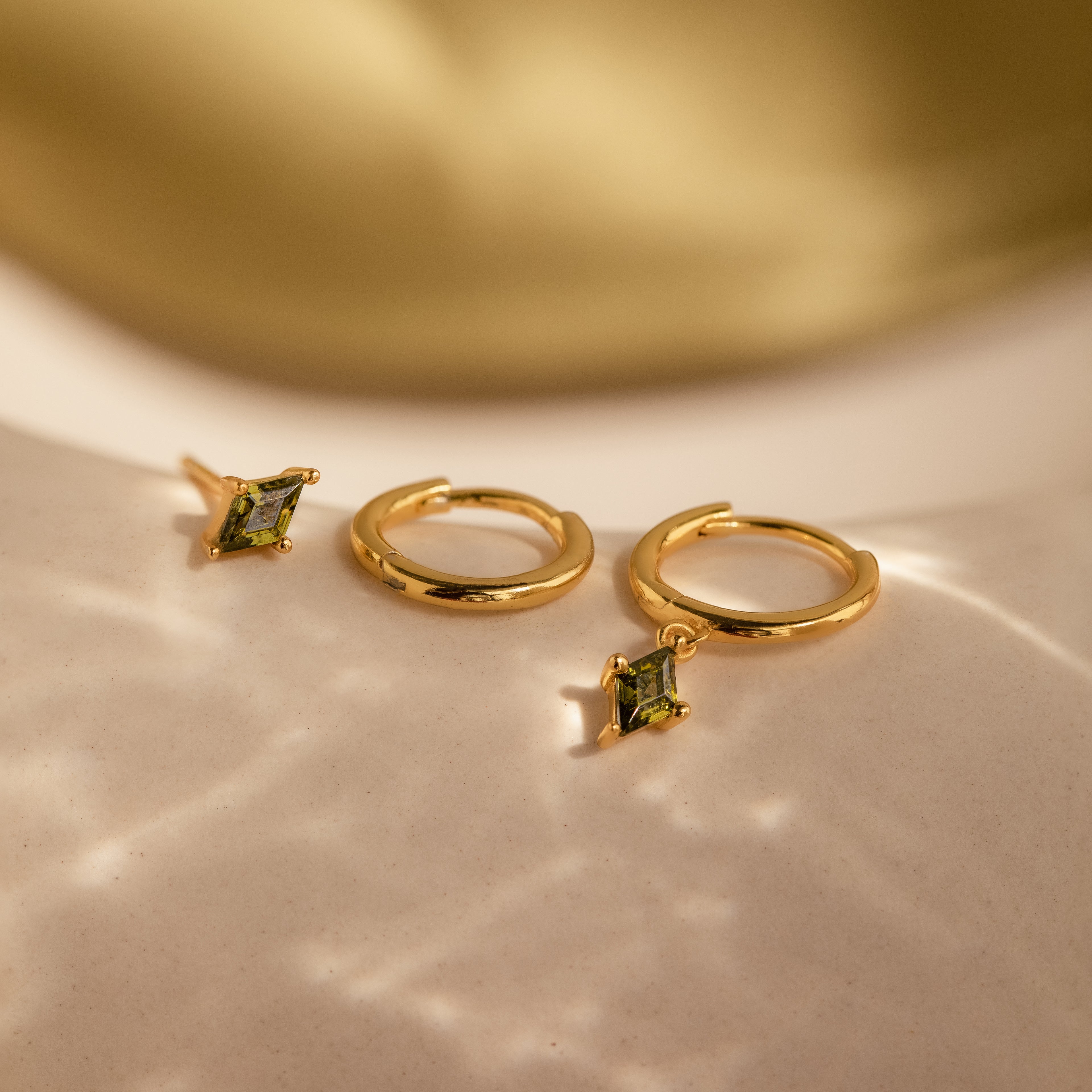The Peridot Earrings Set in 18K Gold features gold hoop stacking earrings with green gemstone charms, beautifully displayed on a creamy, softly lit surface.