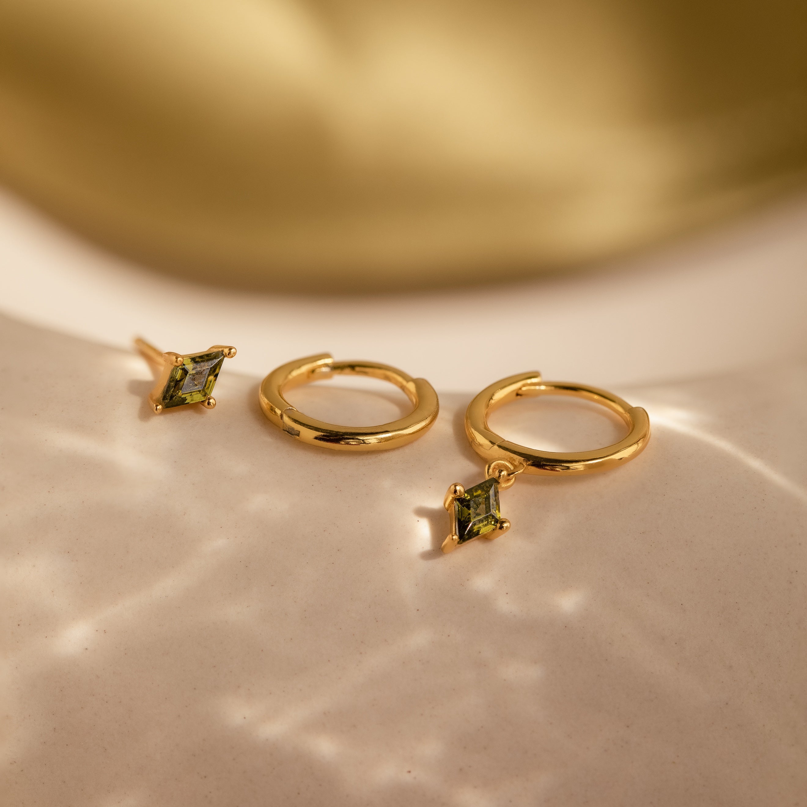 The Peridot Earrings Set features two gold hoops with square peridot gemstone charms, displayed on a light textured surface. Wear them stacked or solo for a vibrant accent.