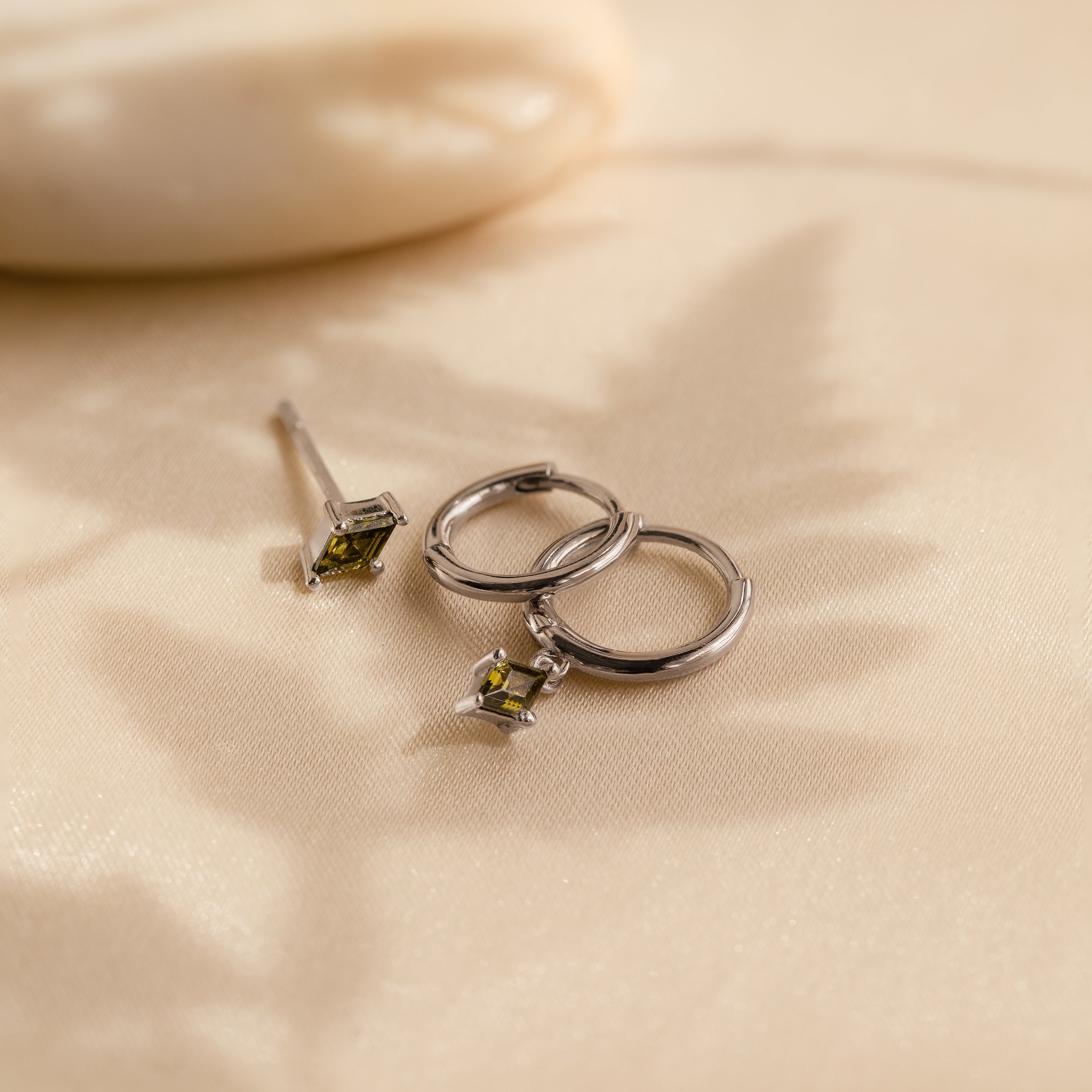 The Peridot Earrings Set in Sterling Silver, featuring two silver stacking earrings and a single peridot earring with green gemstones, rests on beige satin fabric.