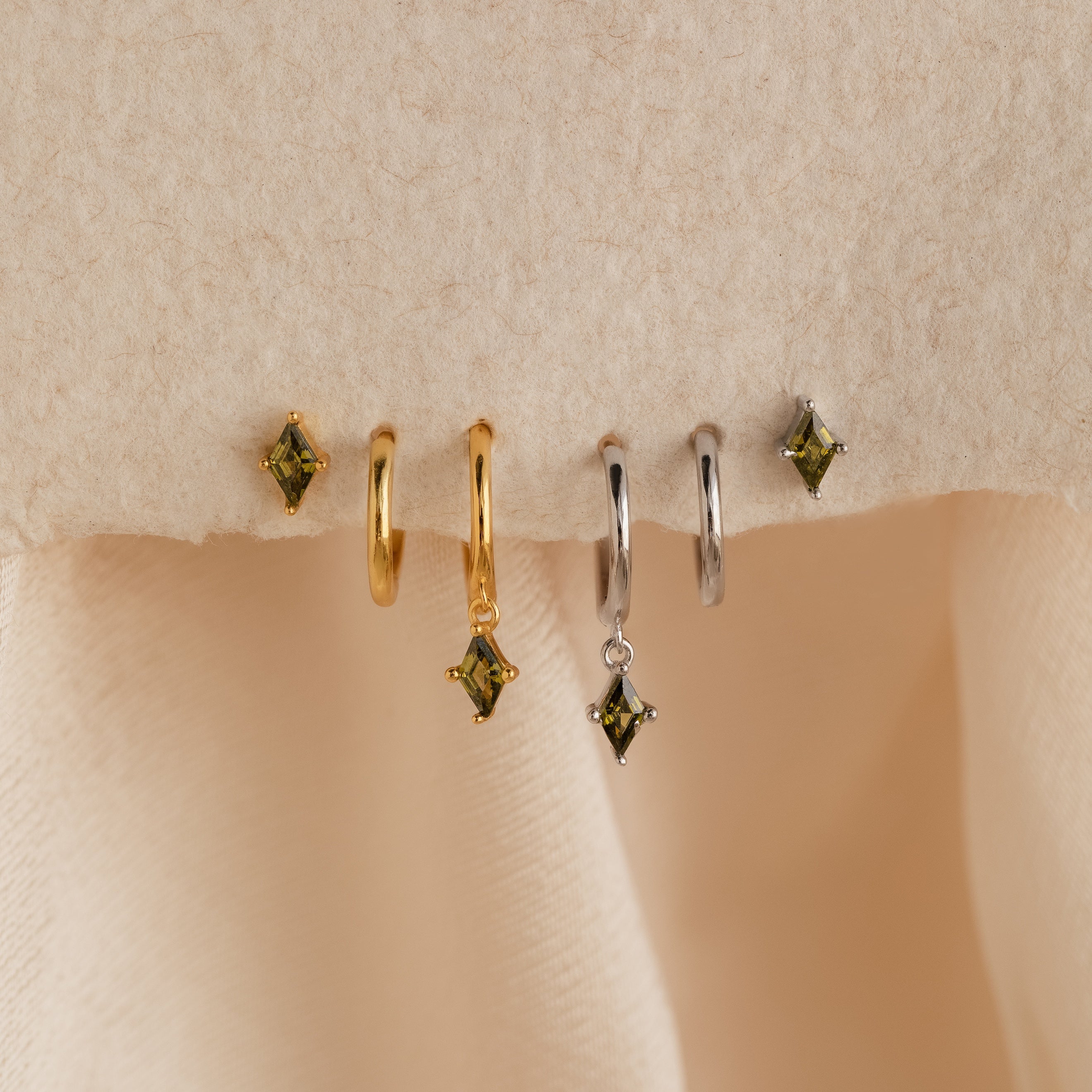 The Peridot Earrings Set includes two pairs of hoop earrings—one gold, one silver—each featuring a green peridot gemstone charm, all displayed on beige fabric. Perfect for fans of stacking earrings.