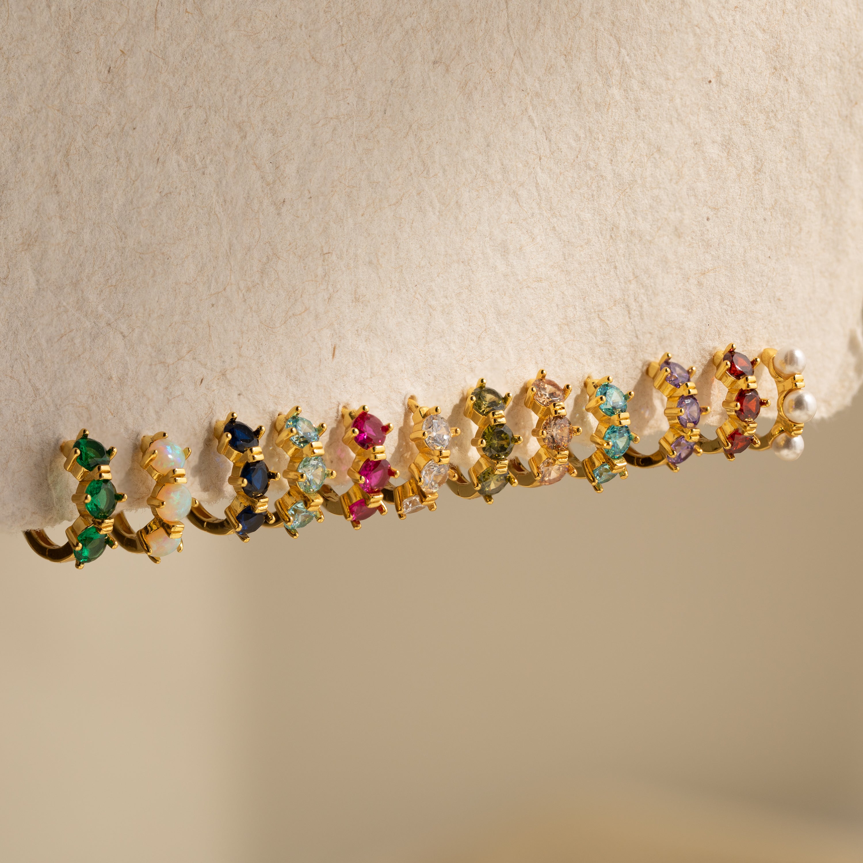 Triple Birthstone Huggies—gold hoop earrings adorned with colorful gemstones and pearls—add a birthstone jewelry charm or create your own custom look. Displayed neatly on beige fabric, they’re perfect for personalizing your style.