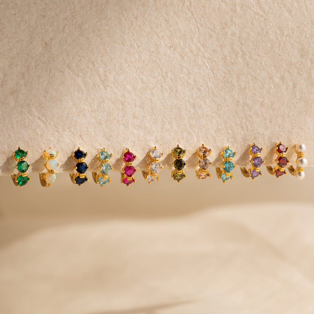 Triple Garnet Huggies, featuring vibrant garnet gemstones in a stylish huggie design, are displayed on a textured beige surface.