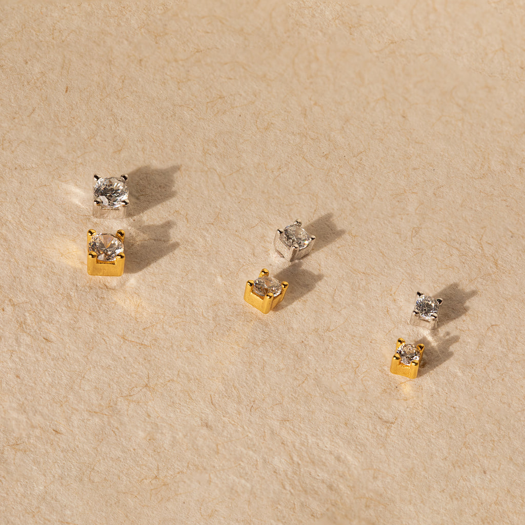 The Diamond Flatback Studs Set in Sterling Silver features three pairs—two gold, one silver—displayed on a textured beige surface. Includes classic square studs with secure screw backs for comfortable, subtle charm.