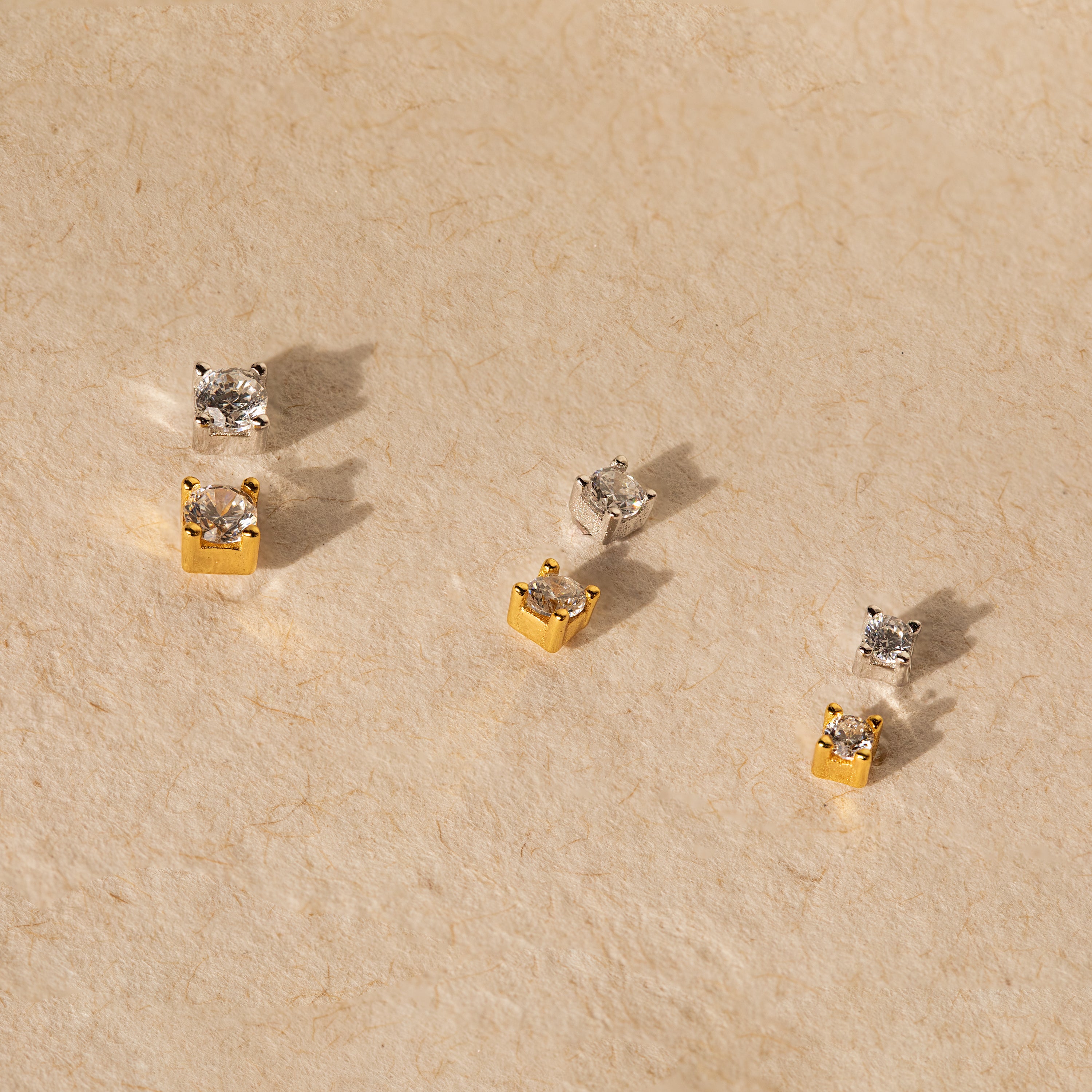 The Diamond Flatback Studs Set in Sterling Silver features three pairs—two gold, one silver—displayed on a textured beige surface. Includes classic square studs with secure screw backs for comfortable, subtle charm.