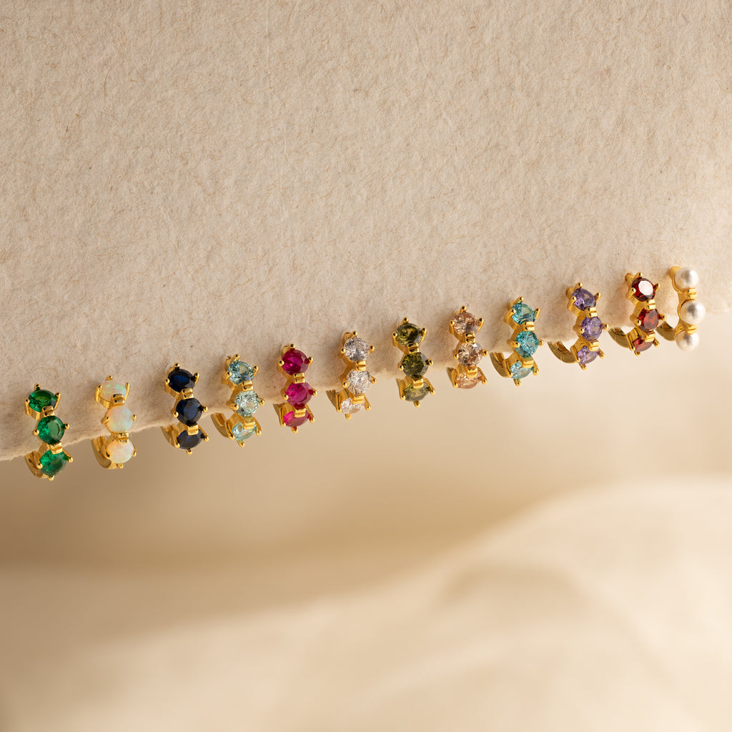 Triple Diamond Huggies displayed on a textured beige surface, featuring minimalist gold settings and sparkling gemstones.