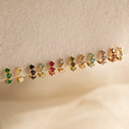 Twelve pairs of small, colorful customizable earrings, including Triple Blue Zircon Huggies, minimalist hoops, and two pearl studs are arranged on a beige background.