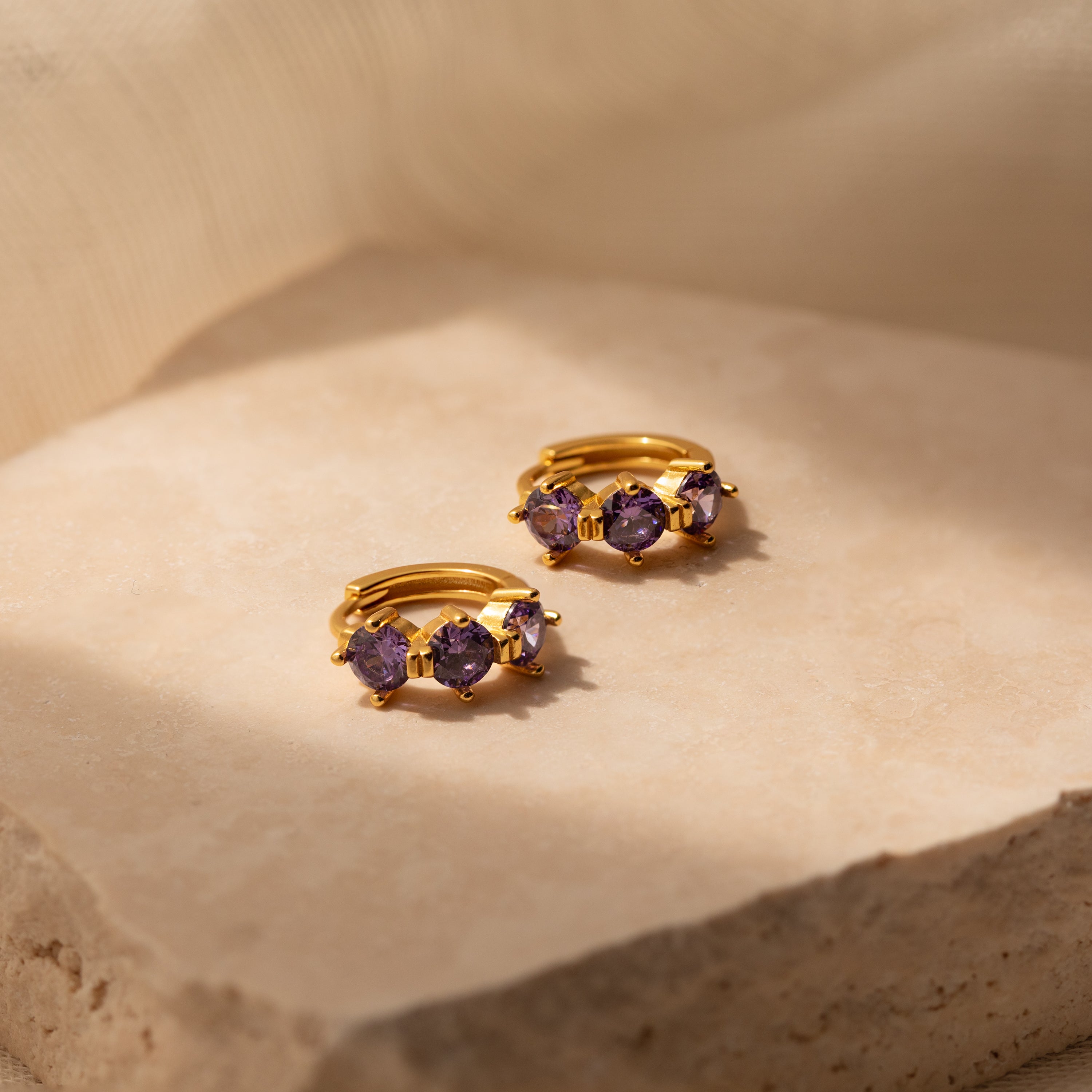 The Triple Amethyst Huggies are gold hoop earrings featuring three purple gemstones each, showcasing minimalist jewelry style and displayed on a beige stone surface in soft lighting.
