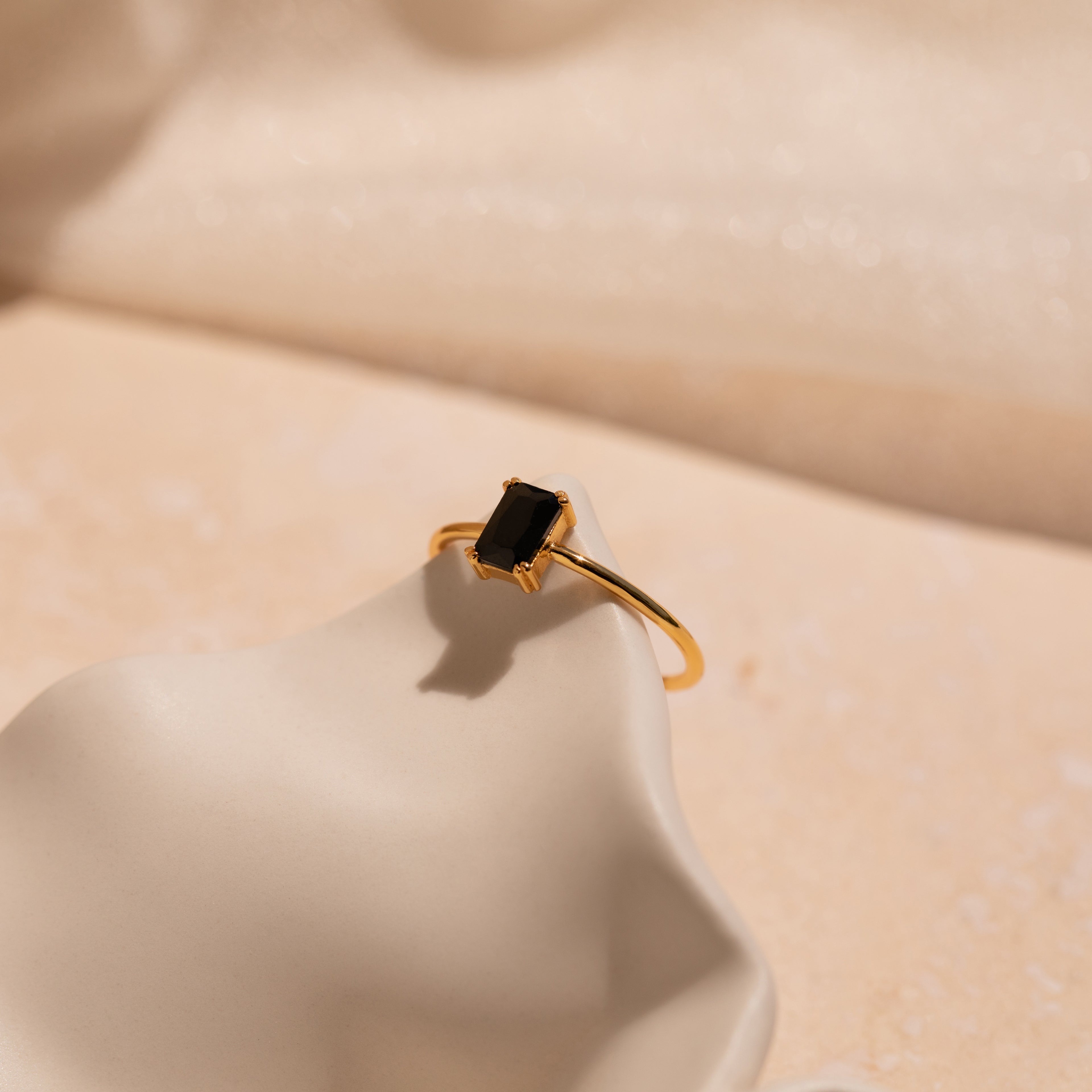 The Black Emerald Ring is a vintage-inspired gold ring featuring a rectangular black gemstone, perfect as an Art Deco piece and shown on a smooth cream-colored surface.