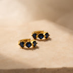 Triple Sapphire Huggies—gold minimalist hoops with three deep blue sapphires, shown on a beige stone in soft light; ideal personalized jewelry for gemstone lovers.
