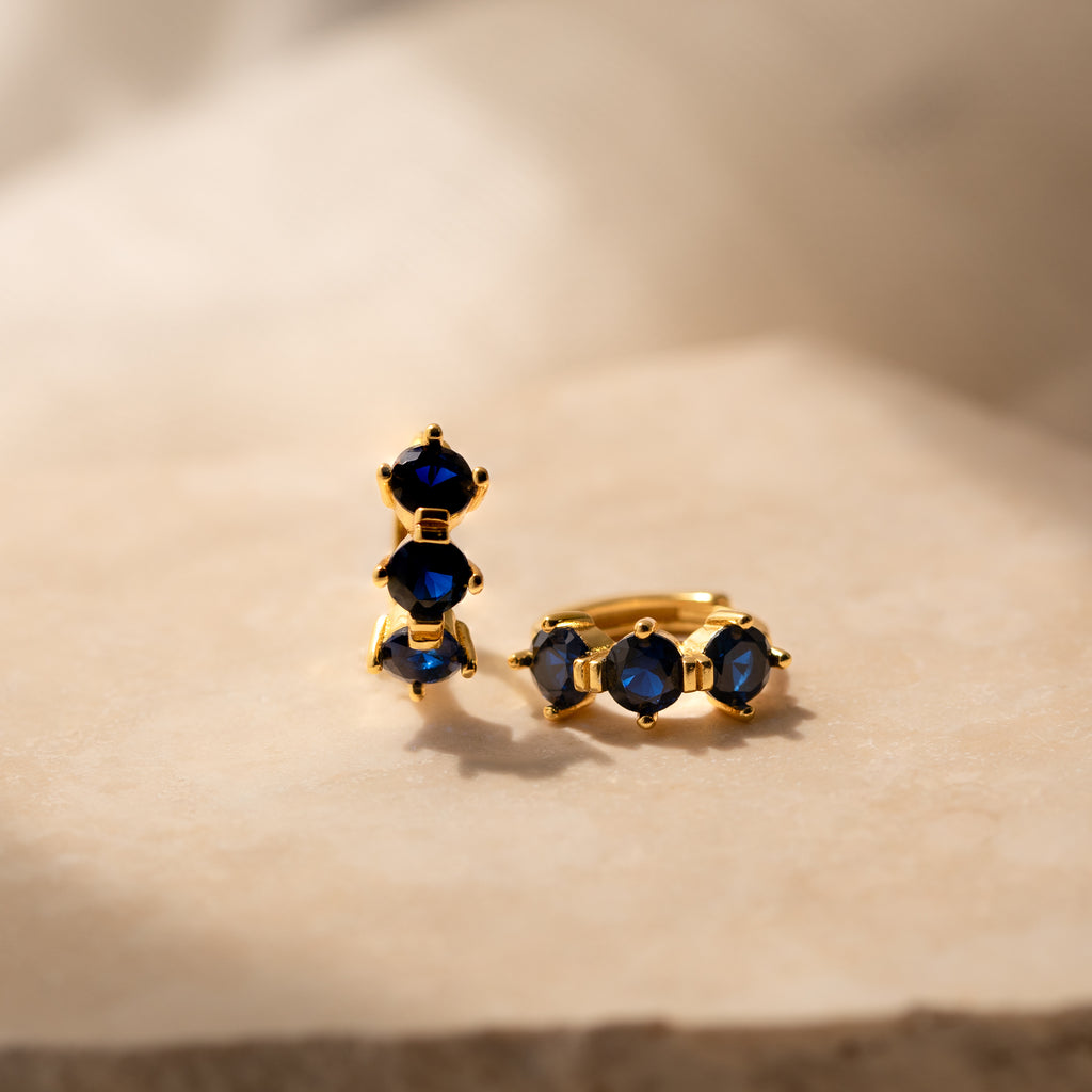 Triple Sapphire Huggies featuring gold earrings with three deep blue sapphires each, displayed on beige marble in soft light—ideal minimalist hoops for those who love personalized jewelry.