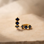 Triple Sapphire Huggies featuring gold earrings with three deep blue sapphires each, displayed on beige marble in soft light—ideal minimalist hoops for those who love personalized jewelry.