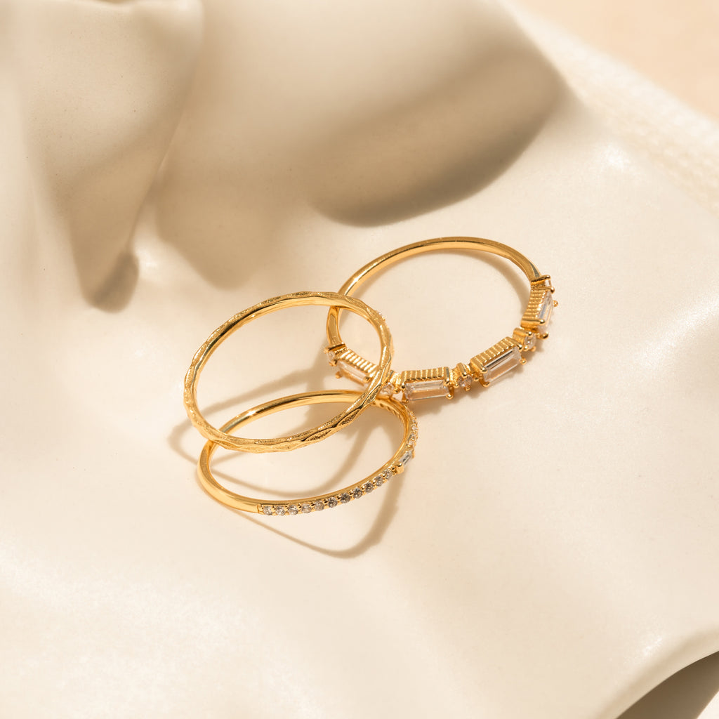 The Diamond Stacking Ring Set, featuring three delicate gold rings including a shimmering diamond band, is displayed on cream-colored satin—making it a perfect Christmas gift.