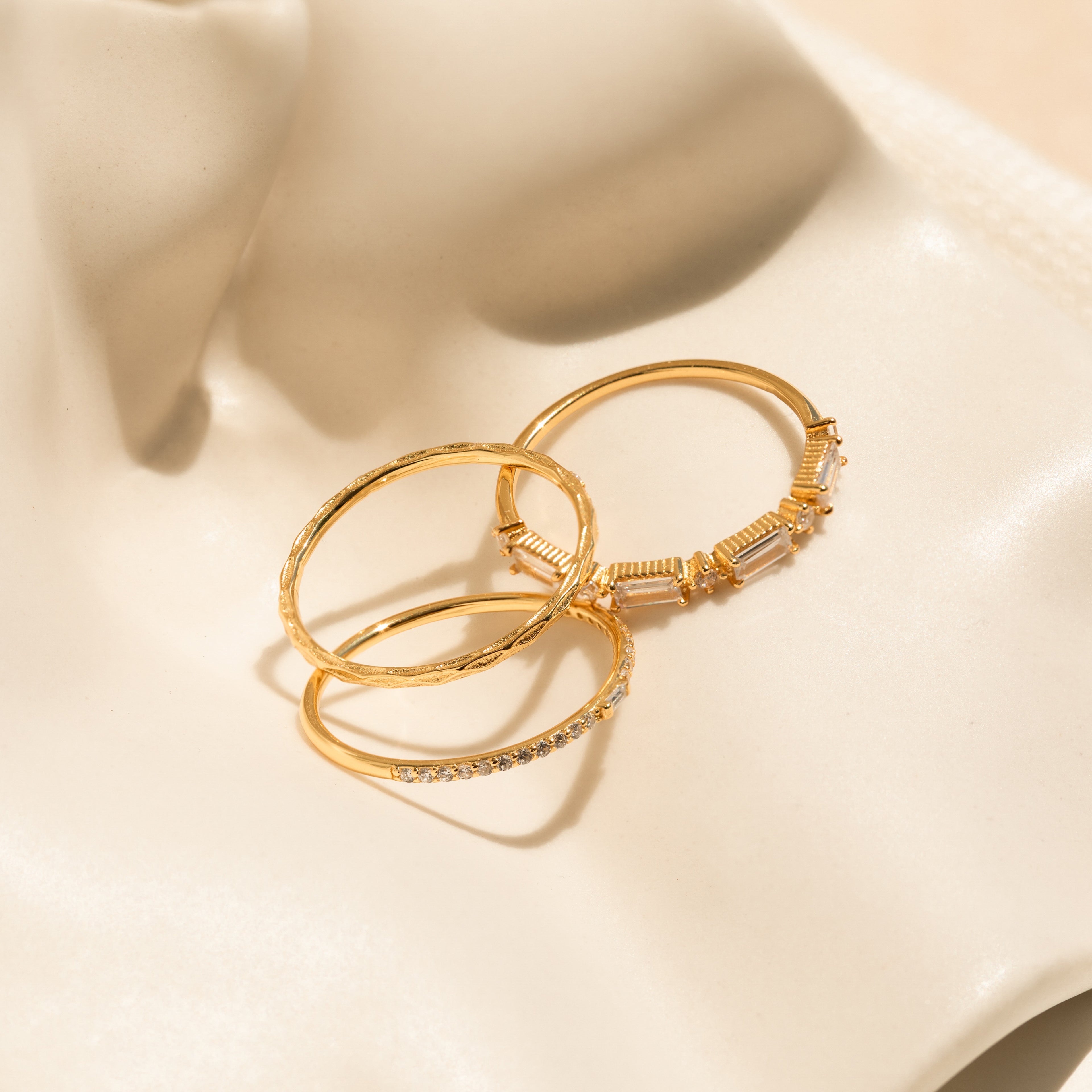 The Diamond Stacking Ring Set, featuring three delicate gold rings including a shimmering diamond band, is displayed on cream-colored satin—making it a perfect Christmas gift.