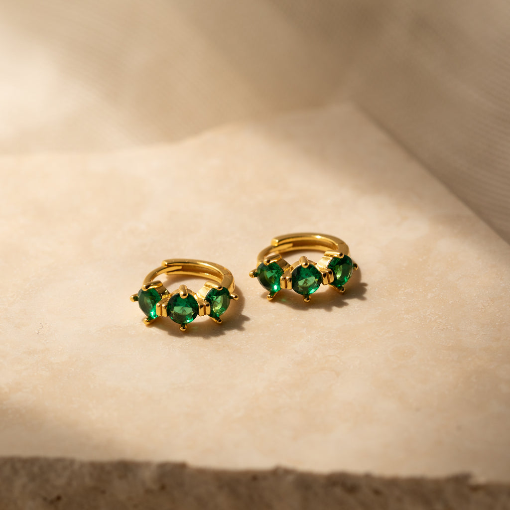 Triple Emerald Huggies: gold hoop earrings with three green gemstones each, shown on a beige textured surface in soft light—perfect personalized jewelry for any occasion.