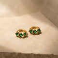 Triple Emerald Huggies: gold hoop earrings with three green gemstones each, shown on a beige textured surface in soft light—perfect personalized jewelry for any occasion.